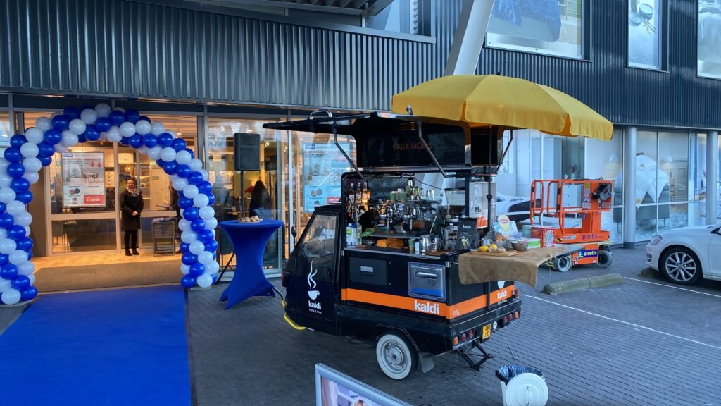 Mobiele koffiebar Piaggio Ape tijdens een bedrijfsevenement met blauwe loper en ballonnen.